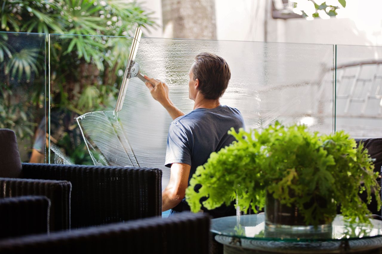 A man cleaning an outdoor glass barrier with a squeegee, with greenery all around.